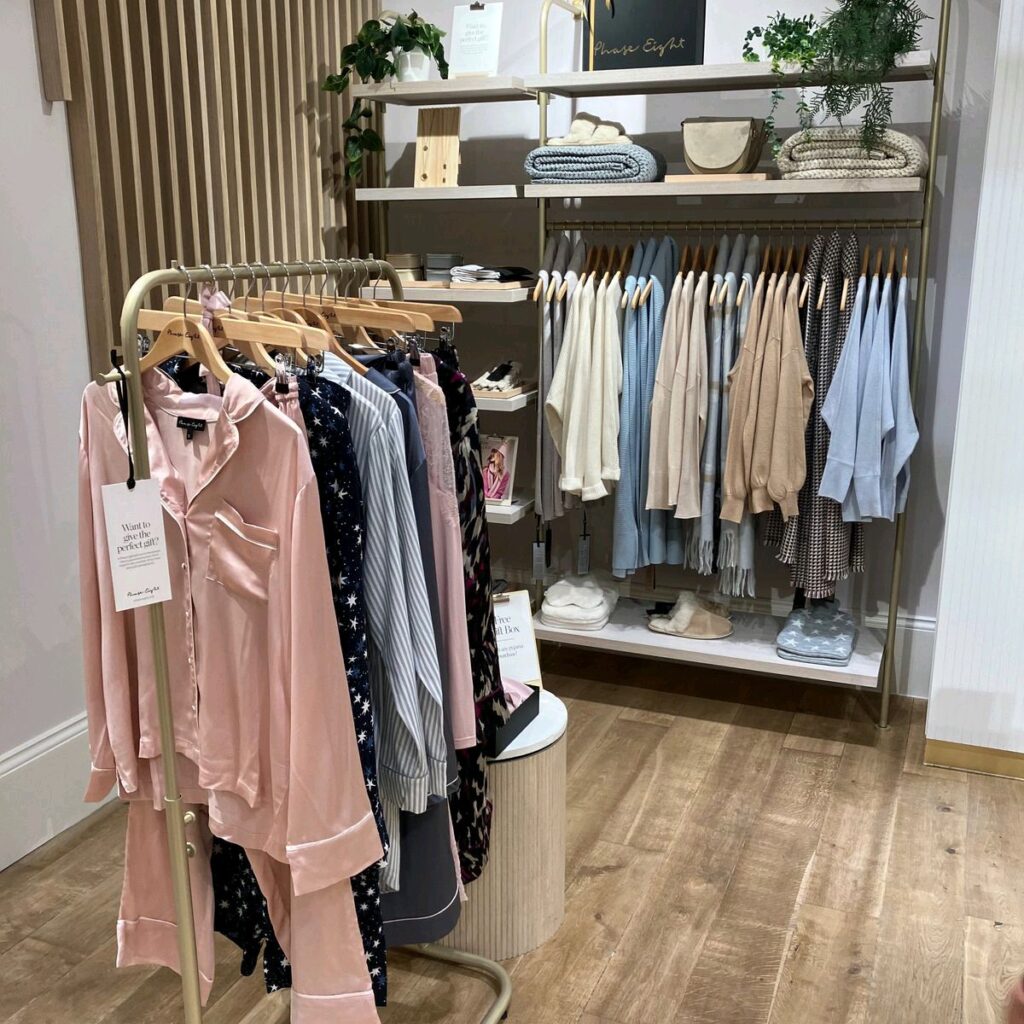 HOL Group A cozy clothing store display featuring a rack of soft pink and patterned pajama sets from Phase Eight in the foreground. Behind, there are shelves and hanging rods showcasing neatly arranged tops and sweaters in neutral tones. The setting includes wooden elements and greenery decor.