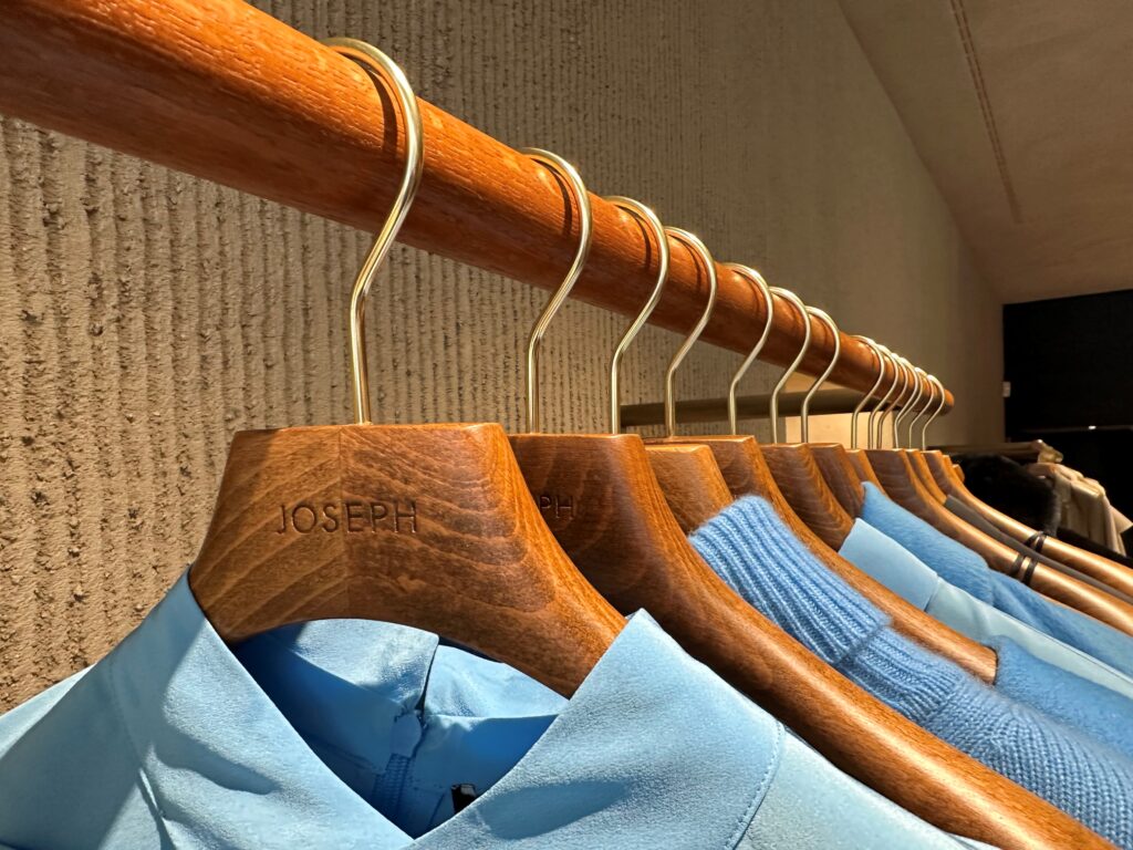 HOL Group A row of wooden hangers, each engraved with the name "Joseph," showcases an assortment of light blue shirts and sweaters on a wooden rod against a textured beige wall. The neatly arranged clothing exudes sophistication in an upscale retail environment.