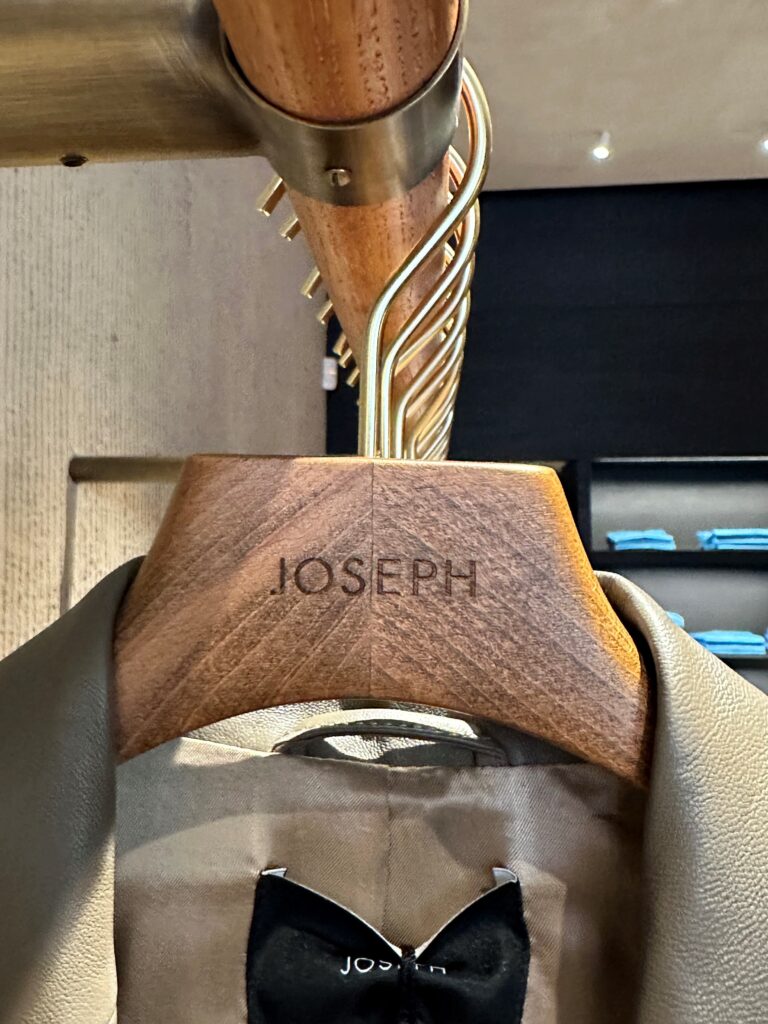 HOL Group A wooden coat hanger engraved with the name "JOSEPH" holds a beige leather jacket on a metal rack. The background is blurred, showcasing shelves filled with neatly folded blue clothing items.
