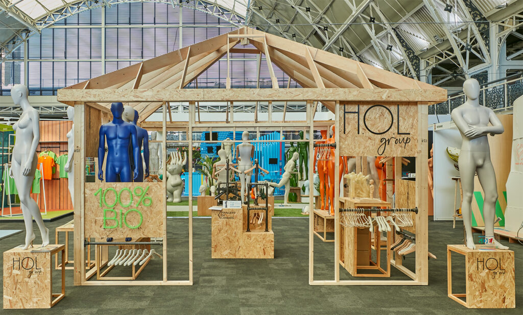 HOL Group At the 2024 VM & Display Show, a modern exhibition booth designed like a wooden house showcases mannequins displaying eco-friendly clothing. Signs read "HOL Group" and "100% BIO." The booth features a mix of colorful and neutral attire, with additional displays and clothing racks within the space.