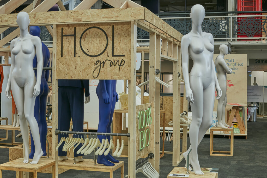 HOL Group A display area at the 2024 VM & Display Show features two white mannequins standing under a wooden structure with the text "HOL group" on it. Nearby are clothing racks with hangers and additional mannequins, as well as various wooden frames and decor elements in an indoor setting.