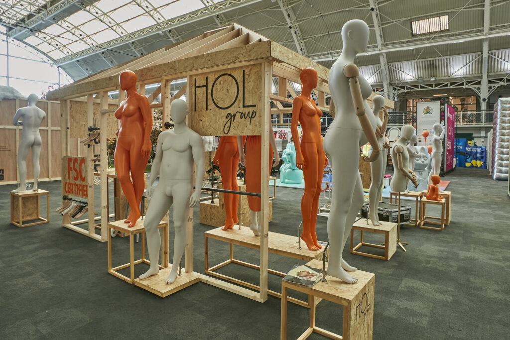 HOL Group A display area inside a spacious, industrial-style exhibition hall featuring numerous mannequins in various poses and colors, primarily white and orange. The mannequins are arranged on and around wooden platforms. Signs on the displays read "HOL group" at the VM & Display Show 2024.
