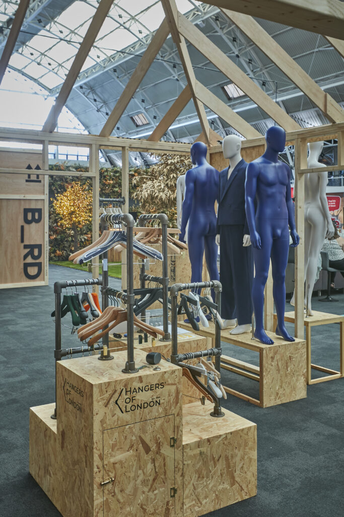 HOL Group An indoor exhibition for Show 2024 features wooden mannequins sporting various clothes, including blue bodysuits and formal attire. Display racks with hangers and accessories stand nearby, all within a structure with a wooden framework and an illuminated, spacious ceiling, showcasing top-tier VM.