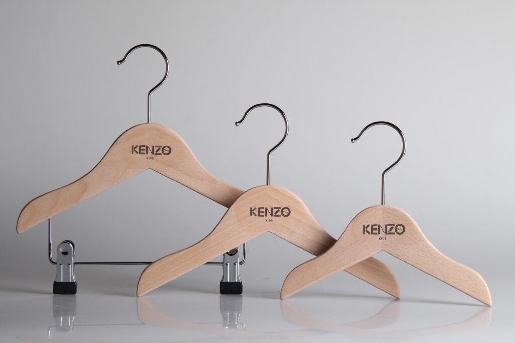 HOL Group Three wooden coat hangers with metal hooks are displayed against a plain background. Each hanger has the brand name "Kenzo" printed on it. One hanger includes clips for hanging pants or skirts.
