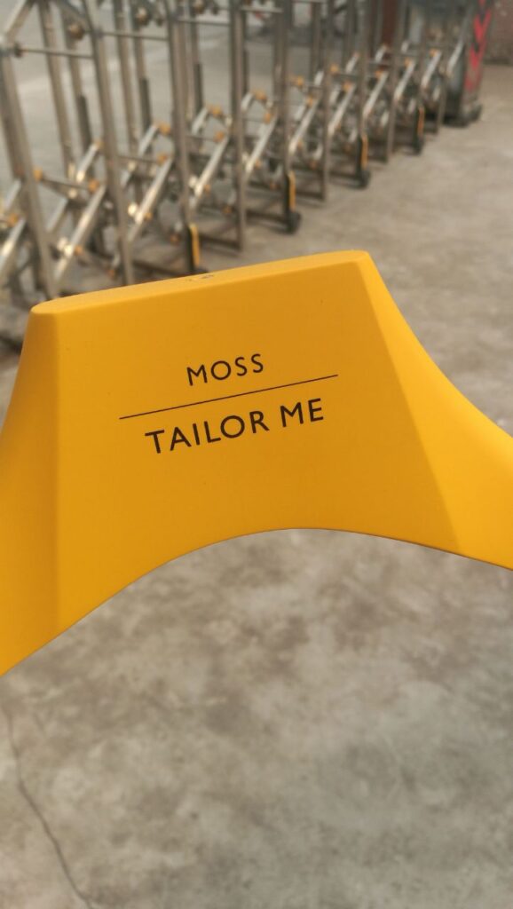 HOL Group A yellow clothing hanger with "MOSS TAILOR ME" elegantly printed on it. In the background, a soft focus unveils metal frames and a concrete floor, reminiscent of the timeless style of Moss Bros.